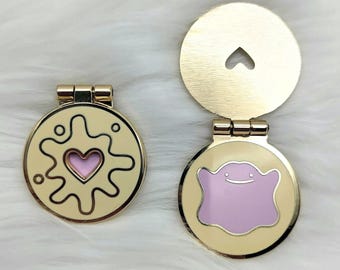 Puede incluir: Dos espejos compactos dorados con bisagras. Uno presenta un diseño de corazón rosa, el otro un personaje rosa Ditto. Los compactos son redondos con un acabado dorado y un recorte en forma de corazón en la tapa.