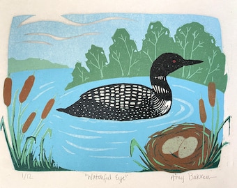May include: A black and white loon with a red eye swims in a blue lake with green trees and brown cattails. The loon is swimming towards a nest with two brown eggs. The text "Watchful Eye" and "Amy Bakken" are written below the image.
