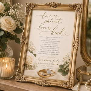 May include: A framed print with the text "Love is patient, love is kind" and a gold ornate frame. The print includes a wedding ring set and floral accents. The frame is displayed on a wooden surface with a candle and flowers.