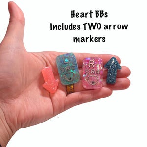 May include: Heart BBs with two arrow markers. The image shows a collection of resin-based accessories. There are two rectangular pieces with the letters "L SRC" and "R SRC" and a heart design. A pink arrow and a blue glitter arrow are also included.