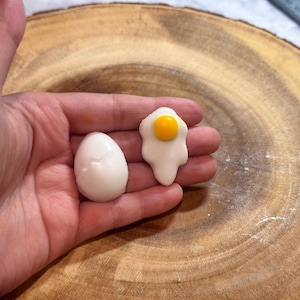 May include: Two white egg-shaped silicone molds, one shaped like a fried egg with a yellow yolk, and the other a whole egg.