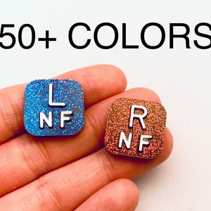 May include: Two square-shaped eye chart stickers with glitter. The left sticker is blue with white letters that say "L NF". The right sticker is orange with white letters that say "R NF".