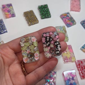 May include: Two rectangular resin pendants are held in a hand, featuring embedded designs. One has a floral and koala bear theme, the other has a star and cartoon character theme. Other resin pendants with various designs are scattered around.