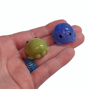 May include: Two small, handmade resin figurines. One is a light green frog with black eyes, and the other is a blue, glittery octopus. The figurines are held in a hand, showcasing their size and detail. The frog and octopus are whimsical and cute.