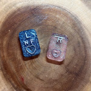 May include: Two small, glittery, rectangular resin charms with silver lettering. The left charm is blue with the letters "LNF" and a silver heart. The right charm is pink with the letter "R" and a silver heart.