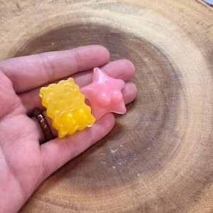 May include: Two small, colorful, handmade soap bars. One is yellow and shaped like a honeycomb. The other is pink and shaped like a star.