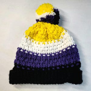 Op de afbeelding: Een gehaakte muts met horizontale strepen in geel, wit, paars en zwart. De muts is afgewerkt met een veelkleurige pompon die alle vier de kleuren bevat. De textuur is gecreëerd door dikke garensteken in een herhalend patroon.