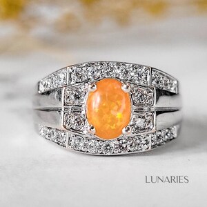 May include: A silver ring with an oval, orange gemstone at the center, surrounded by small, clear stones. The ring has a layered design with the word "LUNARIES" in the bottom right corner.