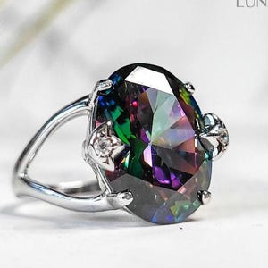 May include: A silver ring featuring a large, oval-cut gemstone with a vibrant, iridescent color display of blues, greens, purples, and yellows. The ring has a split shank design and small accent stones on the sides of the main stone.