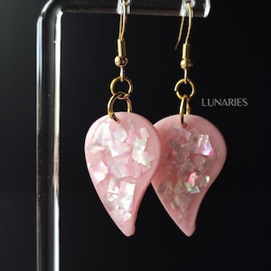May include: A pair of teardrop-shaped earrings in light pink with embedded iridescent flakes. The earrings feature gold-toned metal hooks and jump rings. The word LUNARIES is printed in white text on the right side of the image.