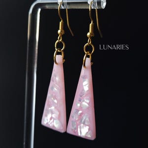May include: A pair of drop earrings featuring triangular pink resin pendants embedded with iridescent white shell fragments. The pendants hang from gold-toned metal fishhook wires. The word LUNARIES is printed in white text on the right side.