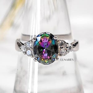 May include: A silver-toned ring featuring a large, oval-cut, iridescent rainbow-coloured centre stone flanked by three small, clear, round-cut stones on each side. The word LUNARIES is printed in small black capital letters below the ring setting.