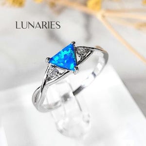 May include: A silver ring with a triangular, blue opal center stone and two smaller clear stones. The ring band has a twisted design. The word "LUNARIES" is in the upper left corner.