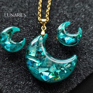 May include: A jewelry set featuring a crescent moon pendant on a gold-tone chain and matching crescent moon stud earrings. Each piece is made of translucent teal resin embedded with iridescent blue and green shell fragments. The text LUNARIES is visible.