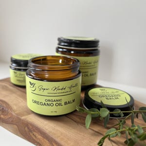 May include: Three amber glass jars with black lids containing organic oregano oil balm. The light green labels feature the text Super Minded Health, the website address, and the product name Organic Oregano Oil Balm with a net weight of 2oz/56g.