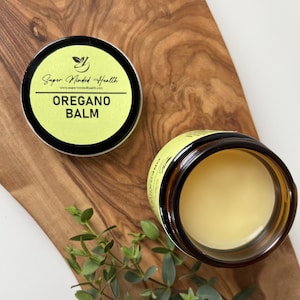 May include: A round amber glass jar containing a pale yellow balm, accompanied by a separate black lid featuring a light green label. The label displays the text Super Minded Health, the website address, and the words OREGANO BALM in bold black letters.
