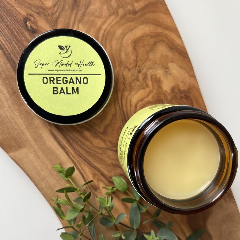 May include: A round amber glass jar containing a pale yellow balm, accompanied by a separate black lid featuring a light green label. The label displays the text Super Minded Health, the website address, and the words OREGANO BALM in bold black letters.
