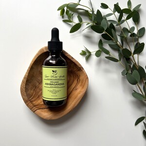 May include: A brown glass bottle with a dropper top containing organic Ashwagandha herbal supplement. The bottle is sitting in a wooden bowl with a green leafy branch in the background.