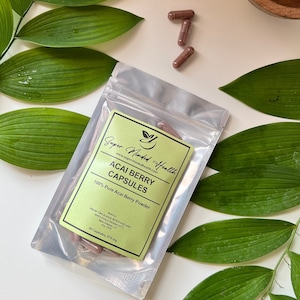 May include: A silver pouch with a green label that reads "Super Minded Health" and "Acai Berry Capsules". The label also states "100% Pure Acai Berry Powder" and "50 Capsules, 575 mg". There are several brown capsules scattered around the pouch.
