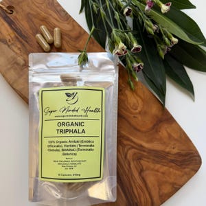 Op de afbeelding: Een doorzichtig hersluitbaar zakje met Organic Triphala-capsules en een lichtgroen etiket. Het etiket bevat de tekst Super Minded Health, Organic Triphala, en vermeldt ingrediënten zoals Amlaki, Haritaki, Bibhitaki, en 50 capsules, 610mg.