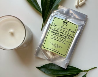 Może przedstawiać: Srebrny worek z białą etykietą, na której widnieje napis "Super Minded Health" i "Organic Apple Cider Vinegar Capsules with the Mother". Worek zawiera białe kapsułki i jest otoczony zielonymi liśćmi.