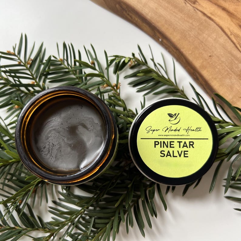 Puede incluir: Un tarro de cristal &aacute;mbar abierto que contiene un b&aacute;lsamo marr&oacute;n oscuro junto a su tapa negra, que presenta una etiqueta amarilla con el texto Super Minded Health, www.supermindedhealth.com y PINE TAR SALVE, sobre agujas de pino verdes.