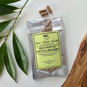 Puede incluir: Una bolsa de plástico transparente que contiene cápsulas marrones etiquetadas como "Wild Crafted Cat's Claw Bark" de Super Minded Health. La etiqueta es verde claro con texto negro. Varias cápsulas están esparcidas alrededor de la bolsa. El fondo es blanco con hojas verdes.