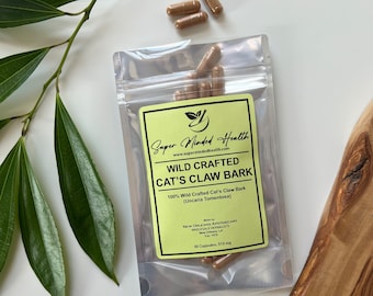 May include: A clear plastic pouch containing brown capsules labeled "Wild Crafted Cat's Claw Bark" from Super Minded Health. The label is light green with black text. Several capsules are scattered around the pouch. The background is white with green leaves.