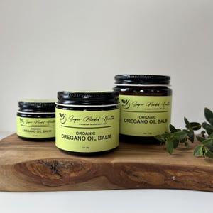 May include: Three amber glass jars with black lids containing Organic Oregano Oil Balm. The labels are light yellow with black text, displaying the brand name Super Minded Health, the website, and net weights of 1 oz, 2 oz, and 4 oz.