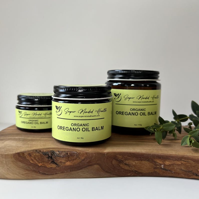 May include: Three amber glass jars with black lids containing Organic Oregano Oil Balm. The labels are light yellow with black text, displaying the brand name Super Minded Health, the website, and net weights of 1 oz, 2 oz, and 4 oz.