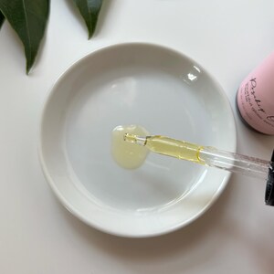May include: A clear glass dropper bottle with a pink label that says "Radiant Glow Serum". A drop of serum is being dispensed from the dropper onto a white ceramic dish.
