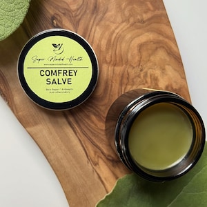 May include: A small, round jar of green comfrey salve with a black label that reads "Super Minded Health" and "Comfrey Salve" with the text "Skin Repair * Antiseptic * Anti-inflammatory" underneath. The jar is sitting on a wooden surface.