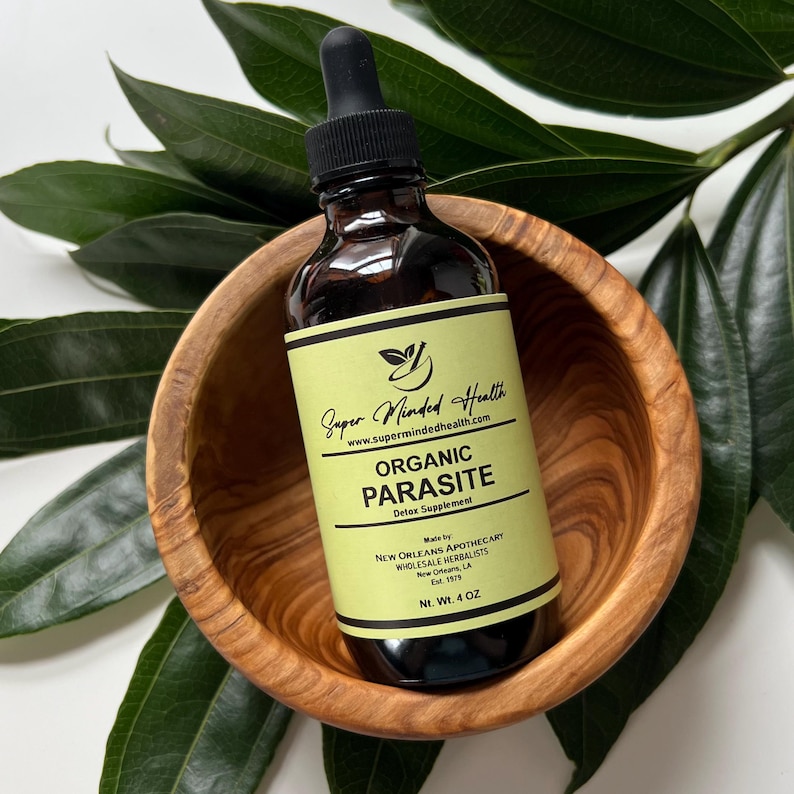 May include: A brown glass bottle with a dropper top, labeled "Super Minded Health Organic Parasite Detox Supplement" with a green and yellow label. The bottle is sitting in a wooden bowl.