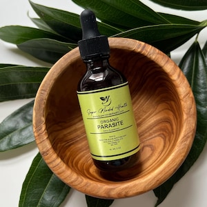 May include: A small, dark brown glass bottle with a dropper top. The bottle is labeled "Super Minded Health" and "Organic Parasite Detox Supplement". The bottle is sitting in a wooden bowl.