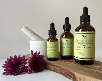 May include: Three amber glass dropper bottles labeled Female Vitality Tonic by Super Minded Health, ranging in size from 1 oz to 4 oz. A white marble mortar and pestle and two dark purple chrysanthemum flowers sit alongside the bottles.