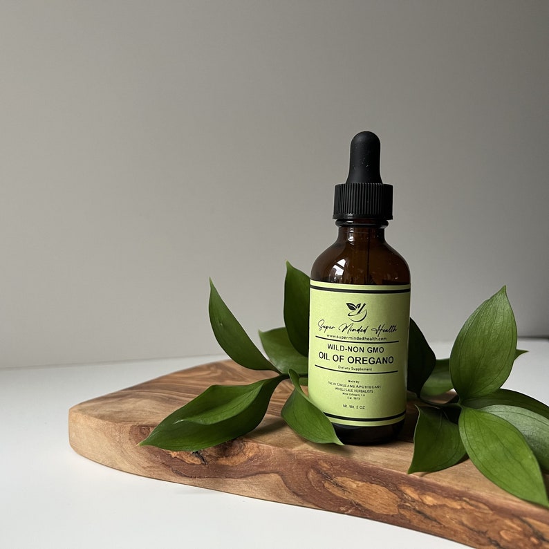 May include: A brown glass bottle with a black dropper top, containing wild-non GMO oil of oregano. The bottle is sitting on a wooden cutting board with green leaves.