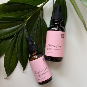 May include: Two amber glass bottles with droppers filled with rosehip oil serum. The bottles have pink labels with black text that reads "Rosehip Oil youthful glow serum Face Oil" and "Net Wt. 1 fl oz / 30 ml" and "Net Wt. 2 fl oz / 60 ml".