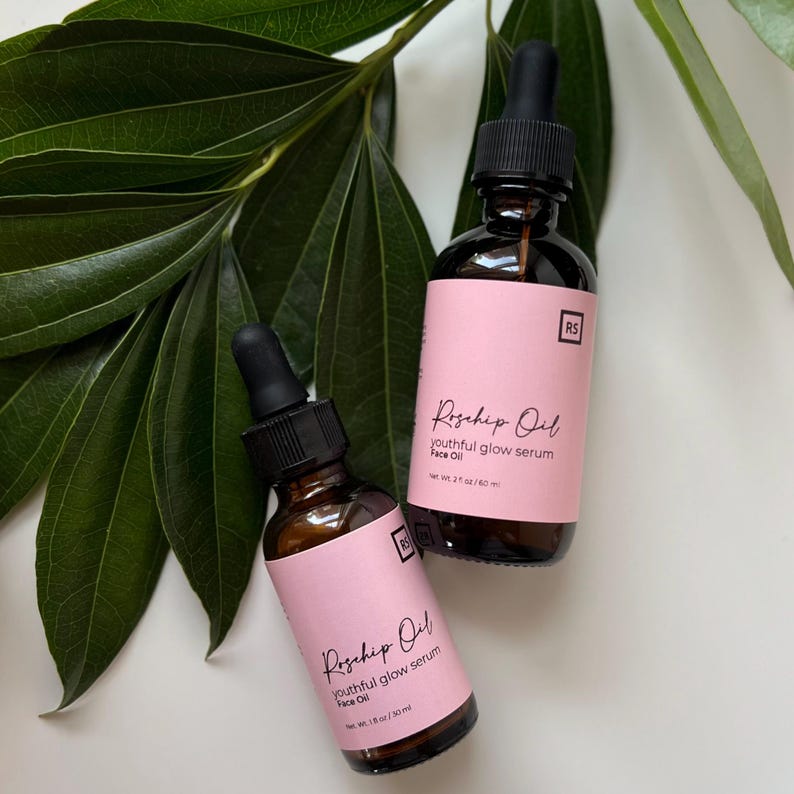 May include: Two amber glass bottles with droppers filled with rosehip oil serum. The bottles have pink labels with black text that reads "Rosehip Oil youthful glow serum Face Oil" and "Net Wt. 1 fl oz / 30 ml" and "Net Wt. 2 fl oz / 60 ml".