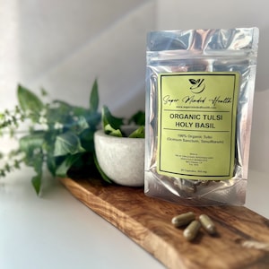May include: A silver pouch containing 50 capsules of organic tulsi holy basil, labeled "Super Minded Health" with the website address "www.supermindedhealth.com". The pouch is on a wooden surface with green basil leaves in a white bowl.