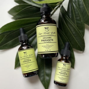 May include: Three brown glass bottles with droppers, labeled "Organic Parasite Detox Supplement" by "Super Minded Health". The bottles are sitting on a white surface with green leaves in the background.
