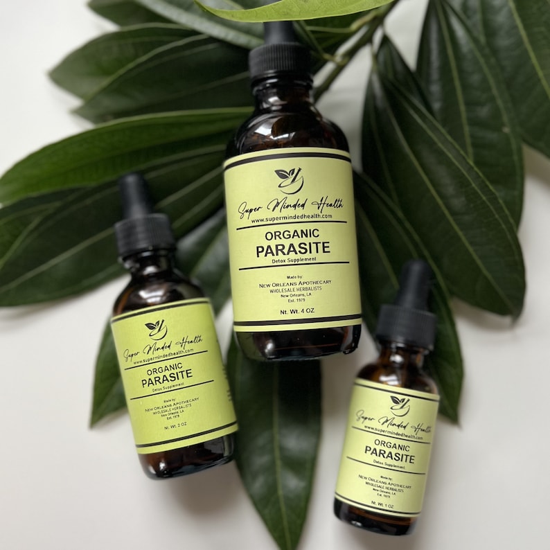 May include: Three brown glass bottles with droppers, labeled "Organic Parasite Detox Supplement" by "Super Minded Health". The bottles are sitting on a white surface with green leaves in the background.