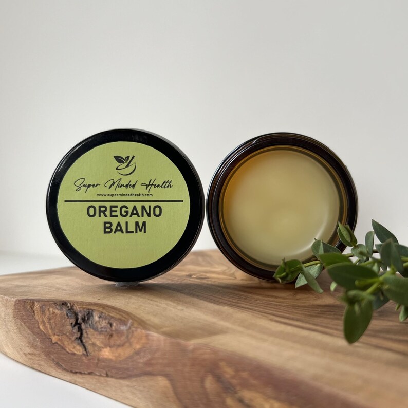 May include: A round container of Oregano Balm with a black lid and a light green label. The label features the text Super Minded Health, the website address, and the words OREGANO BALM. Beside it, an open container reveals a smooth, pale balm.