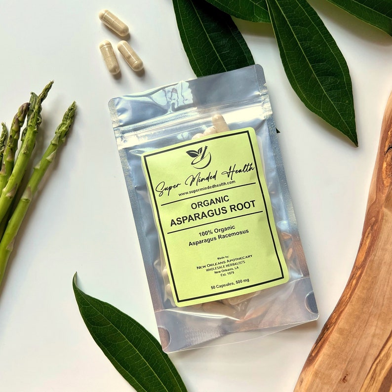 May include: A clear plastic bag with a yellow label that reads "Super Minded Health" and "Organic Asparagus Root". The label also includes the text "100% Organic Asparagus Racemosus" and "50 Capsules, 500 mg". The bag contains capsules and is surrounded by green leaves and asparagus spears.