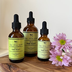 Könnte beinhalten: Drei bernsteinfarbene Glasflaschen mit Tropfern, die Wild-Yamswurzelextrakt enthalten. Die Flaschen sind mit "Super Minded Health" und "Wild Yam Root" sowie der Website-Adresse "www.supermindedhealth.com" beschriftet. Die Flaschen stehen auf einer Holzoberfläche mit rosa Blumen.