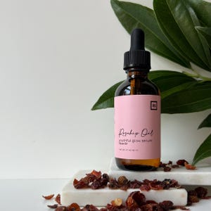 May include: A glass bottle of rosehip oil face serum with a dropper. The bottle is labelled "Rosehip Oil Youthful Glow Serum Face Oil" and has a pink label with black text. The bottle is sitting on a white surface with dried rosehips.