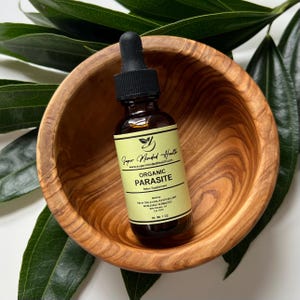 May include: A glass bottle with a dropper top containing a yellow liquid. The bottle is labeled "Super Minded Health" and "Organic Parasite Detox Supplement". The bottle is sitting in a wooden bowl.