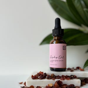 May include: A glass bottle with a dropper top filled with rosehip oil serum. The bottle has a pink label with the text "Rosehip Oil youthful glow serum Face Oil 1 fl oz / 30 ml" and the initials "RS". The bottle is sitting on a white surface with dried rosehips scattered around it.