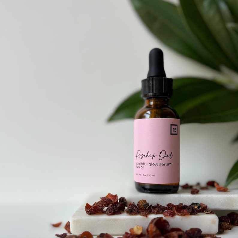 May include: A glass bottle with a dropper top filled with rosehip oil serum. The bottle has a pink label with the text "Rosehip Oil youthful glow serum Face Oil 1 fl oz / 30 ml" and the initials "RS". The bottle is sitting on a white surface with dried rosehips scattered around it.