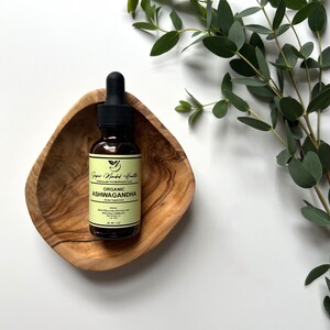 May include: A brown glass bottle with a dropper top containing organic Ashwagandha herbal supplement. The bottle is labelled "Super Minded Health" and "Organic Ashwagandha". The bottle is sitting in a wooden bowl.