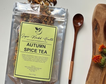 Peut inclure: Un sachet jaune et blanc de thé aux épices d'automne avec une étiquette qui indique "Super Minded Health" et "Autumn Spice Tea". L'étiquette liste les ingrédients, qui comprennent des pommes Honey Crisp biologiques, de l'écorce de cannelle biologique, de l'anis étoilé biologique, de la cardamome biologique, des clous de girofle biologiques, des racines de gingembre biologiques, de la muscade biologique, du zeste d'orange biologique, des pétales de calendula biologiques et du thé rooibos biologique. Le sachet est posé sur une surface blanche à côté d'une cuillère en bois.